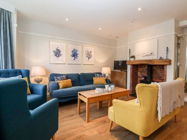 A living room with a blue sofa, yellow armchair, wooden coffee table, fireplace, TV, and framed art on the wall at Fawcett Cottage in Aldeburgh