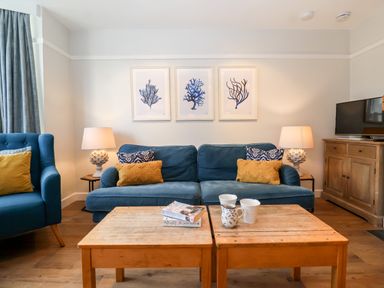 A living room with blue sofa and armchair yellow and patterned cushions wooden coffee tables and framed artworks at Fawcett Cottage in Aldeburgh