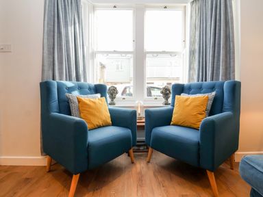 Two blue armchairs with yellow and patterned cushions in front of a window with grey curtains at Fawcett Cottage in Aldeburgh