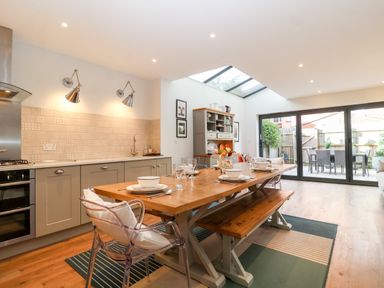 A kitchen dining area with a wooden table set with dishes and glassware a bench and clear chairs light grey cabinets and a tiled backsplash a hutch and sliding glass doors leading to an outdoor dining area at Fawcett Cottage in Aldeburgh