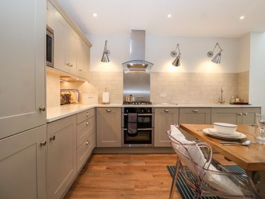 A kitchen with beige cabinets a stove a wooden dining table and transparent chairs at Fawcett Cottage in Aldeburgh