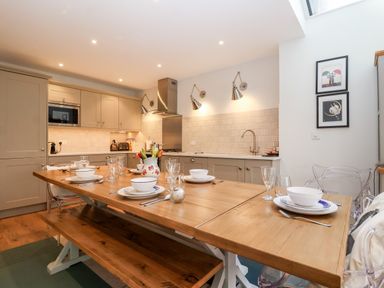 A kitchen with beige cabinets a wooden dining table set with plates bowls glasses and a floral jug at Fawcett Cottage in Aldeburgh