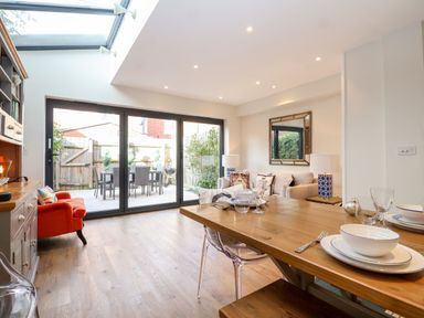 A dining area with a wooden table set with dishes and glasses next to a living area with a beige couch and lamps near glass doors leading to an outdoor patio at Fawcett Cottage in Aldeburgh