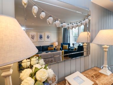 A wooden table with a lamp and flowers in front of a mirror reflecting a living room with blue sofas and framed artwork at Fawcett Cottage in Aldeburgh