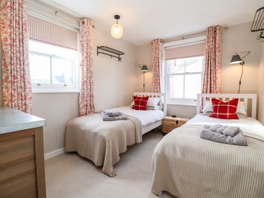 A bedroom with two single beds with beige blankets and red pillows floral curtains and wooden furniture at Fawcett Cottage in Aldeburgh