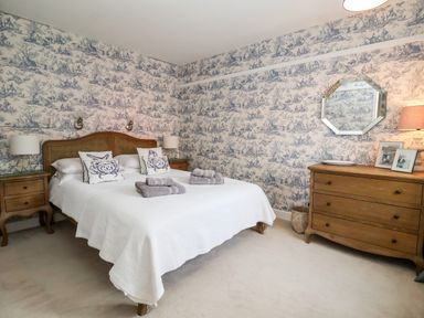 A bedroom with a bed with white bedding and pillows next to wooden nightstands and a wooden dresser with a mirror on the wall at Fawcett Cottage in Aldeburgh