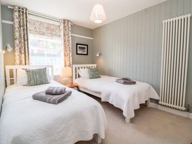 A bedroom with two single beds white bedspreads floral curtains a wall radiator and a bedside lamp at Fawcett Cottage in Aldeburgh