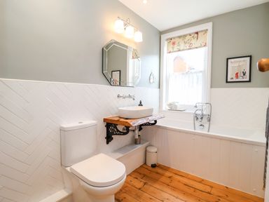 A bathroom with a toilet sink on a wooden shelf a bathtub and a window with floral blind at Fawcett Cottage in Aldeburgh
