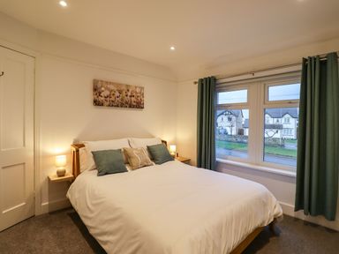 A bedroom with a bed with pillows bedside lamps a window with curtains and a painting above the bed at Maywood in Seahouses