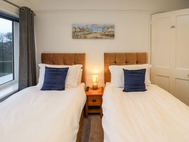A bedroom with two single beds with brown headboards a wooden nightstand with a lamp and a remote control between the beds and a window with dark curtains at Maywood in Seahouses