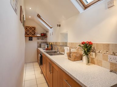 A narrow kitchen with wooden cabinets white countertops and tiled floor with a sink kettle basket and flowers on the countertop at Lythe Cottage in Lythe