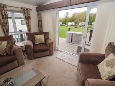 A living room with brown armchairs and a coffee table looking out to a garden through sliding glass doors at Lakeside Haven in Carnforth