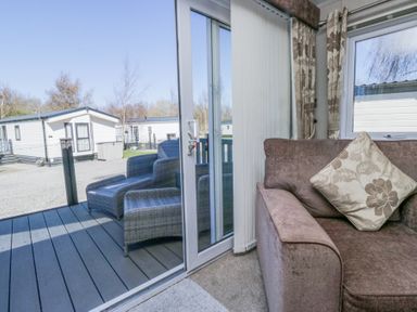 A sofa with a patterned cushion next to sliding glass doors leading to a patio with two wicker chairs at Lakeside Haven in Carnforth