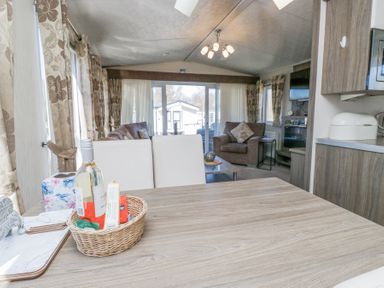 An open living space with a wooden table, white chairs, a basket with wine and snacks, sofas, an armchair, curtains, and a wall-mounted television at Lakeside Haven in Carnforth