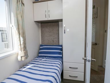 A small bedroom with a single bed with striped bedding a window with curtains overhead cabinets a wardrobe and a door at Lakeside Haven in Carnforth