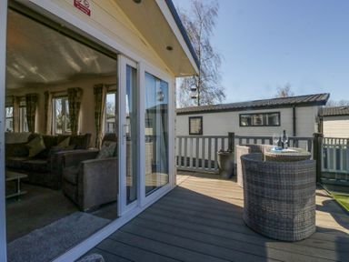 A deck with two wicker chairs and a small table with wine glasses outside a living room at Lakeside Haven in Carnforth