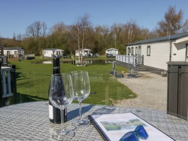 A table with a wine bottle, two wine glasses, a menu, and sunglasses overlooking a green area with cabins and a pond at Lakeside Haven in Carnforth