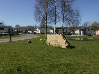 A grassy area with a large stone sign that reads Yealand Conyers near a pond and several modern lodges at Lakeside Haven in Carnforth