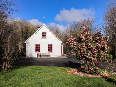 A white house with red window frames and benches outside surrounded by trees and a flowering bush in grass at Kyleatunna Cottage in Ennis County Clare