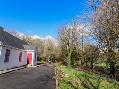 A white house with red windows and door next to a driveway and trees at Kyleatunna Cottage in Ennis County Clare