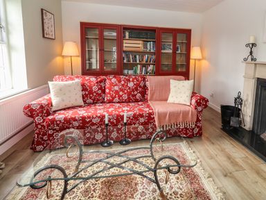 A living room with a red floral sofa two cream cushions pink blanket a glass coffee table on a patterned rug a red bookcase with books two floor lamps and a fireplace at Kyleatunna Cottage in Ennis County Clare