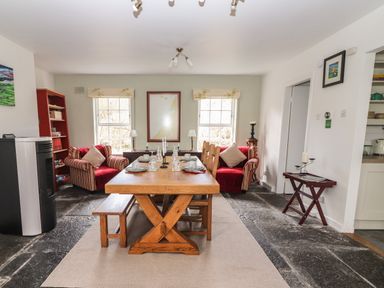 A dining room with a wooden dining table set with plates and glasses two red armchairs by two windows and a bookshelf at Kyleatunna Cottage in Ennis County Clare