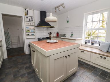 A kitchen with a central island and tiled countertop a stove and range hood a sink under a window with plants and cushions on a bench at Kyleatunna Cottage in Ennis County Clare