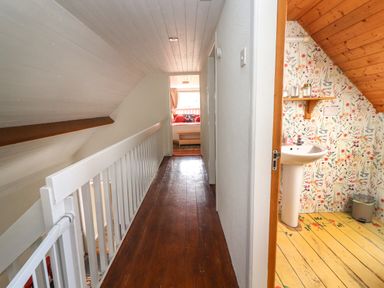 A hallway with wooden floor and white railing leading to a room with a window and a sink area with floral wallpaper and yellow floor at Kyleatunna Cottage in Ennis County Clare