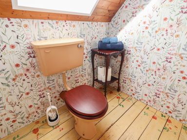 A small bathroom with a beige toilet and wooden seat a wooden floor with floral designs and floral wallpaper at Kyleatunna Cottage Ennis County Clare