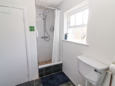 A bathroom with a shower area with tiled walls and curtain a toilet a window with bottles on the sill and a door at Kyleatunna Cottage in Ennis County Clare
