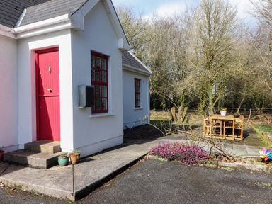 A white cottage with a red door and window frame potted plants on a path and a wooden table with chairs on a patio surrounded by trees at Kyleatunna Cottage in Ennis County Clare