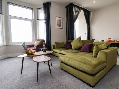 A living room with a green sectional sofa and a purple armchair near large windows with black curtains at Apt 3 @ Hunter's Quay in Bridlington