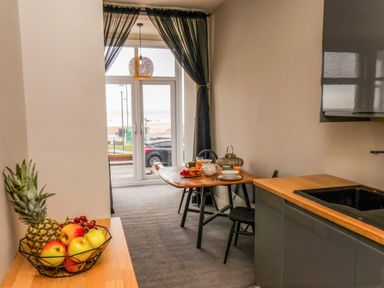 A kitchen area with a fruit bowl on a counter and a small dining table set with breakfast items near a window with black curtains at Apt 3 @ Hunter's Quay in Bridlington