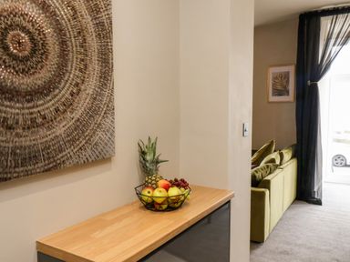 A countertop with a fruit basket holding apples grapes and a pineapple next to a wall with circular artwork a living area with a green sofa and sheer black curtains at Apt 3 @ Hunter's Quay in Bridlington