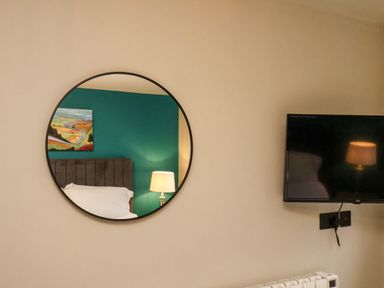 A round wall mirror reflecting a bed with white bedding a lamp and a colorful painting next to a mounted flat screen tv in a room at Apt 3 @ Hunter's Quay in Bridlington