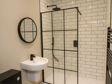 A bathroom with a wall-mounted sink a round mirror and a glass shower enclosure with black grid pattern at Apt 3 @ Hunter's Quay in Bridlington