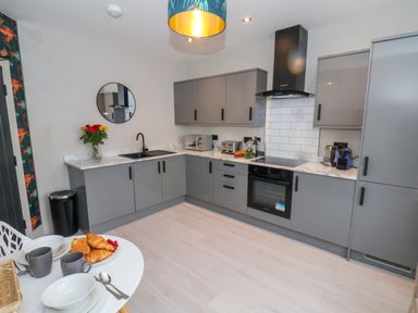 A kitchen with gray cabinets a round mirror flowers on the counter and a dining table with croissants and cups at Apt 6 @ Hunter's Quay in Bridlington