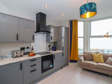 A kitchen with grey cabinets and appliances next to a beige sofa with pillows by windows with yellow curtains at Apt 6 @ Hunter's Quay in Bridlington