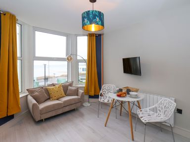 A small living room with a beige sofa orange curtains a round white table with two white chairs and a wall-mounted TV at Apt 6 @ Hunter's Quay in Bridlington
