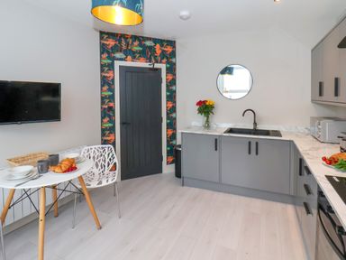 A kitchen with a small dining table with plates and cups a wall-mounted television a gray door with floral wallpaper and gray cabinets at Apt 6 @ Hunter's Quay in Bridlington