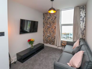 A living room with a gray sofa pink cushions a black media unit with flowers and a wall mounted tv at Apt 6 @ Hunter's Quay in Bridlington