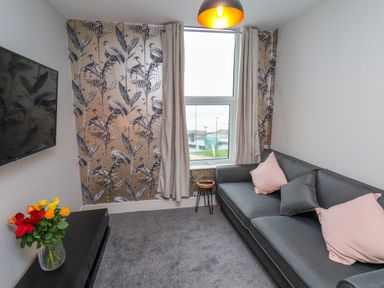 A living room with a gray sofa with three cushions a small round side table a wall-mounted TV colorful flowers in a vase and a window with curtains at Apt 6 @ Hunter's Quay in Bridlington