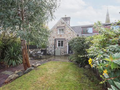 A garden with grass trees and bushes and stone house with patio and outdoor table at 5 The Crescent in Caernarfon