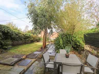 A garden with a stone patio featuring a table and chairs and surrounding trees and shrubs at 5 The Crescent in Caernarfon