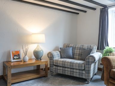 A sitting area with a plaid armchair two cushions a wooden side table with a lamp a framed picture and a vase at 5 The Crescent in Caernarfon