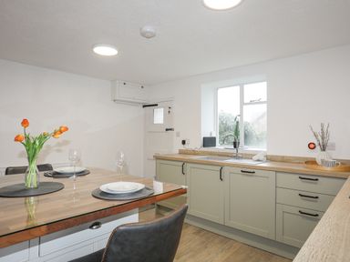 A kitchen with a wooden dining table set with plates and glasses a vase with orange flowers a window above the sink and cabinets at 5 The Crescent in Caernarfon