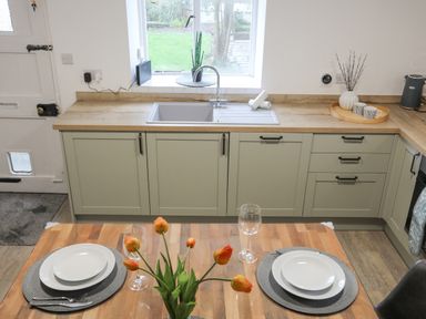 A kitchen with green cabinetry a wooden countertop a sink under a window and a dining table with plates glasses and orange flowers at 5 The Crescent in Caernarfon