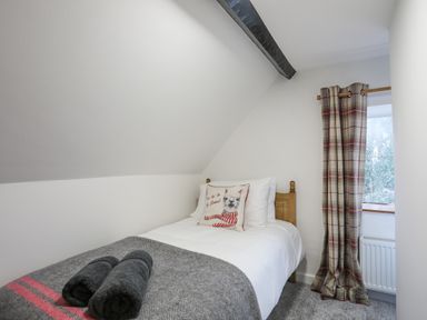 A small bedroom with a single bed a gray blanket two rolled towels a pillow with an animal print and a window with plaid curtains at 5 The Crescent in Caernarfon