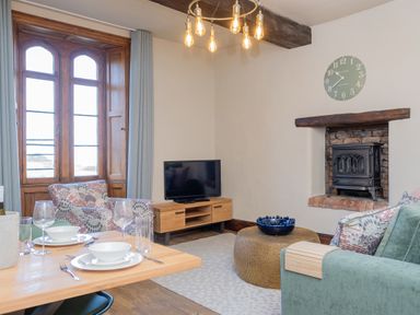 A living room with wooden dining table set for two near a patterned armchair and green sofa a wooden TV stand with a television and a fireplace with a wood stove at Watermouth Castle West Tower Apartment Berrynarbor near Ilfracombe