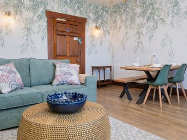 A living room with a green sofa and patterned cushions a round gold coffee table and a wooden dining table with green chairs at Watermouth Castle West Tower Apartment in Berrynarbor near Ilfracombe